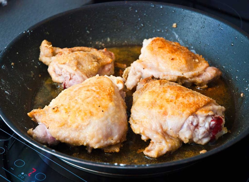 Four seared chicken thighs frying in a pan. 