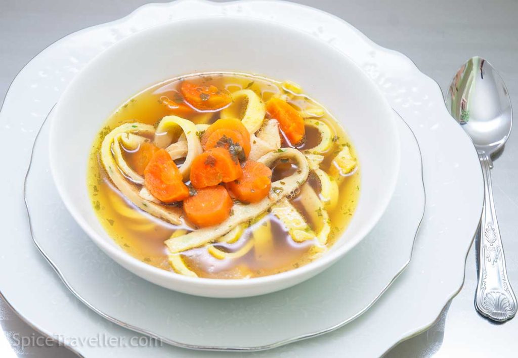 White bowl of pancake strips in Tafelspitz broth, decorated with carrots, presented in an elegant dinner setting.