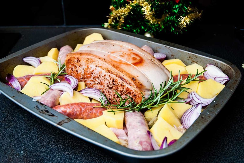 Roasting tray with layer of sauerkraut, potatoes, onions, sausages and boneless pork leg joint in the centre, ready to roast.