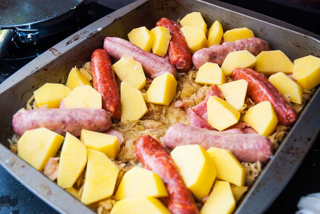 Roasting tray with layer of sauerkraut topped with chunks of potato and two types of sausages before roasting.