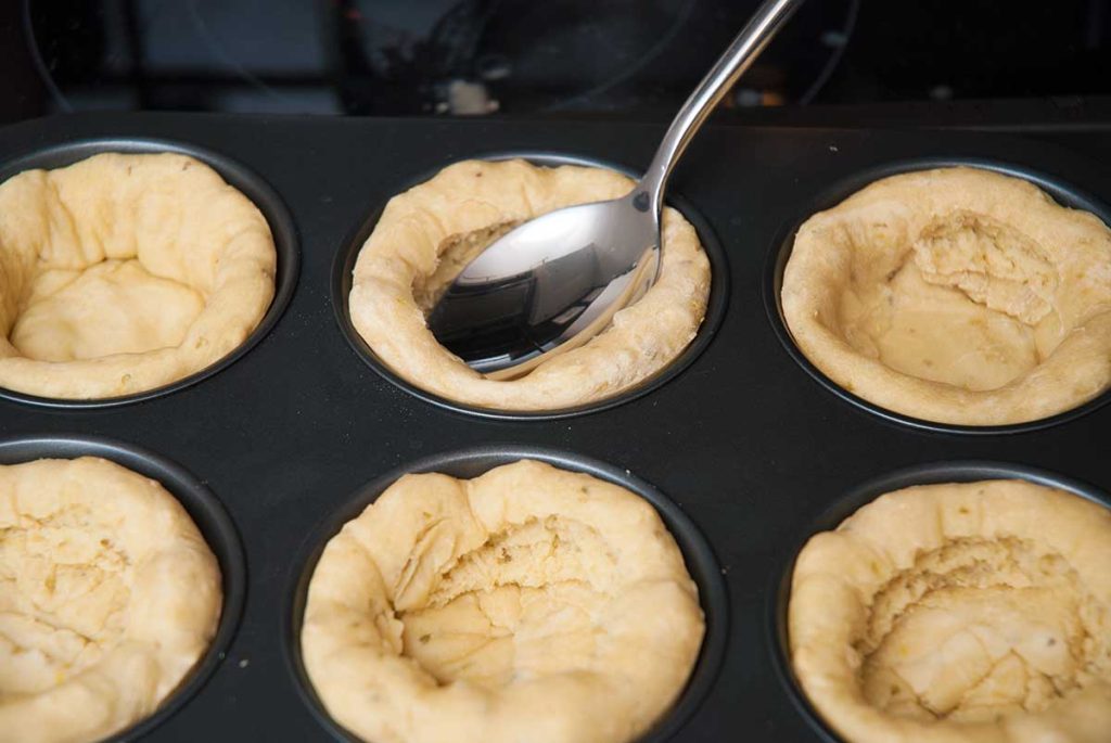 Prebaked mini pizza dough in muffin tin, gently flattened with a teaspoon before adding toppings.