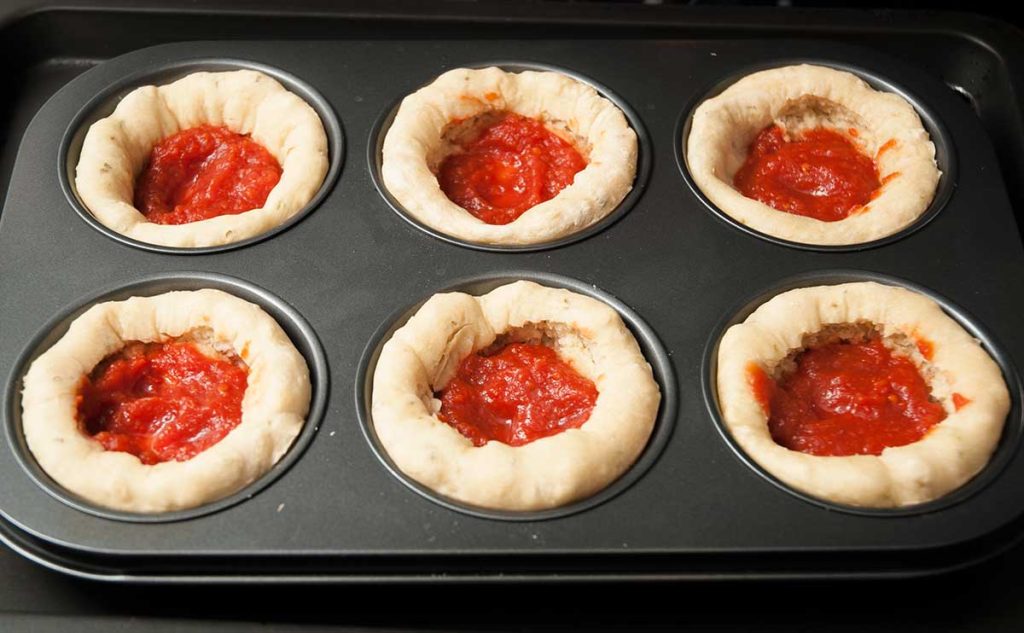 Pizza cupcakes in muffin tin, topped with tomato sauce, ready for the next layer of toppings.