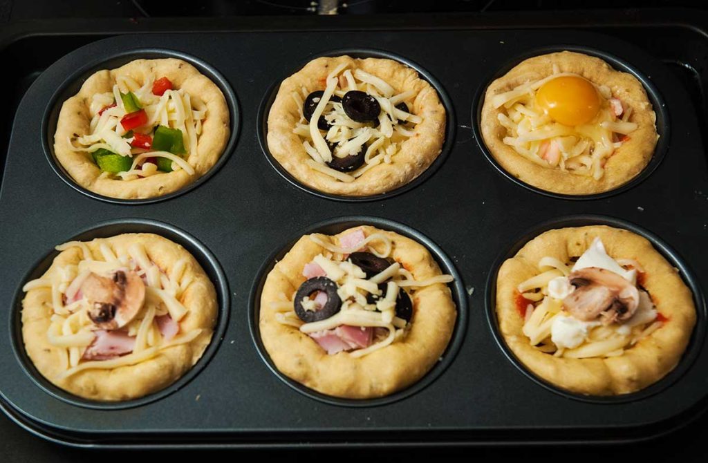 Mini pizza cupcakes filled with  various toppings ready to bake in a muffin tray.