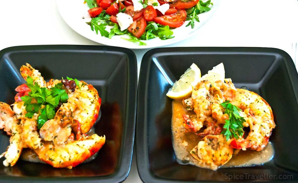 Serving suggestion - two squared black bowls with flambeed langoustine and prawn halved tails in rich aromatic sauce, with lemon wedges and a plate of wild rocket, and tomato salad.