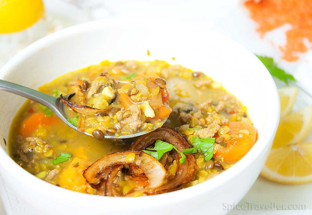 Close-up of a spoon lifting Emirati lamb and lentil soup from a bowl, garnished with lemon slices and fresh parsley, showcasing the soup's rich texture and vibrant ingredients.