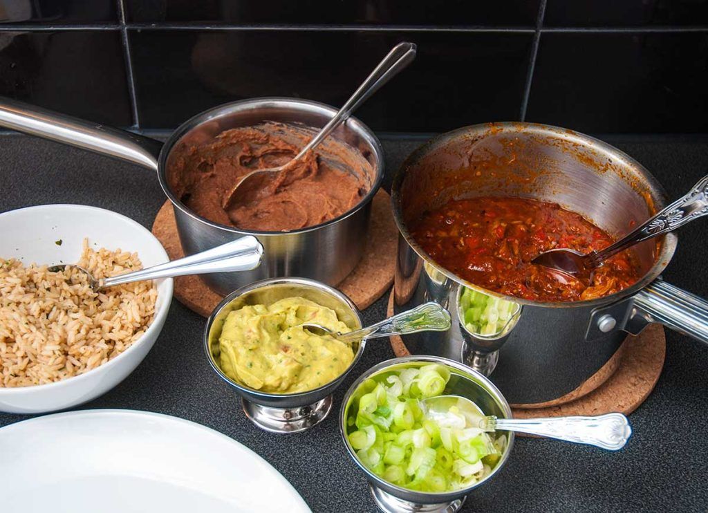All burrito components prepared in separate bowls and pots: refried beans, pork filling, guacamole, rice, spring onions, and tortillas ready for assembly.