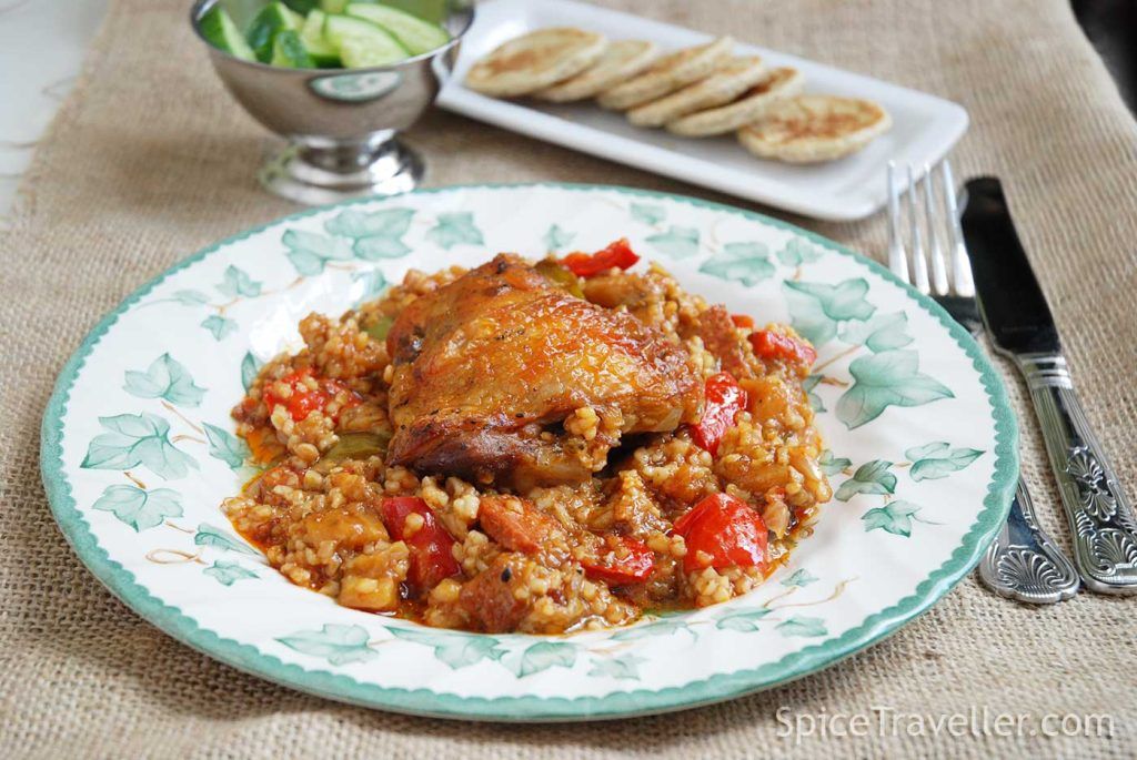 Elegantly plated chicken güveç with a chicken thigh served over bulgur and vegetables, accompanied by sliced cucumber and a separate plate of mini pittas.