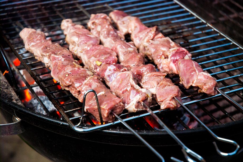 Raw lamb pieces threaded onto skewers, just placed on a charcoal BBQ grill, with visible flames and uncooked meat.