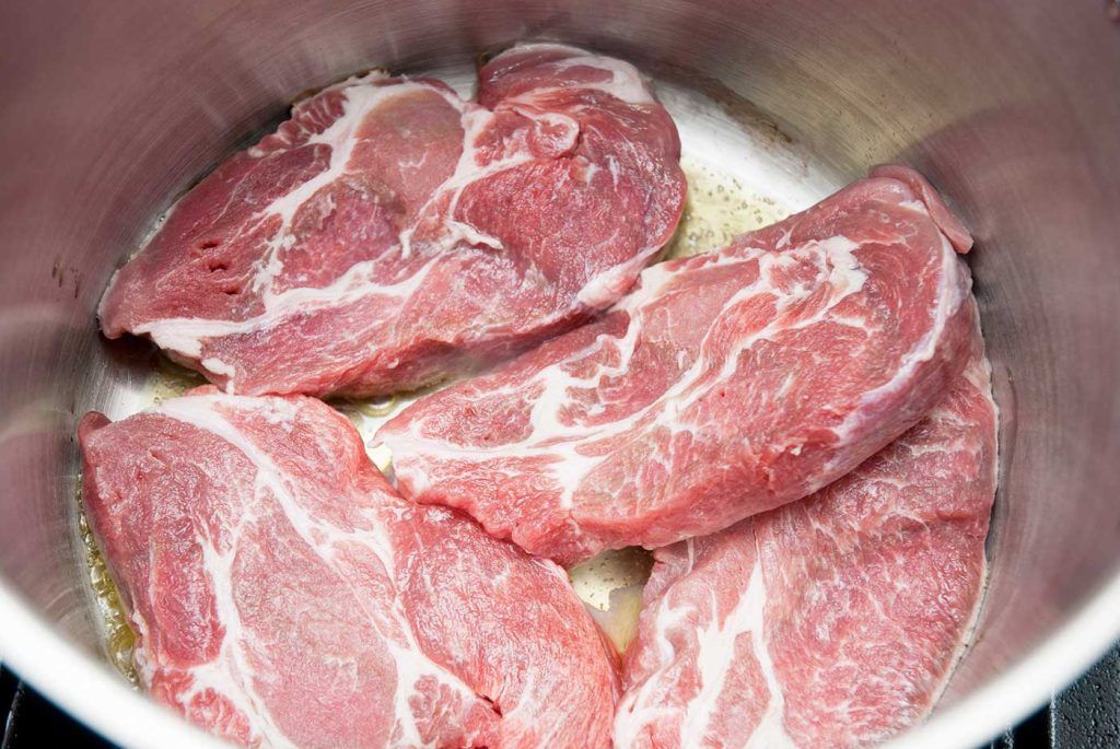 Raw pork shoulder steaks just added to a hot pot, beginning to sear.