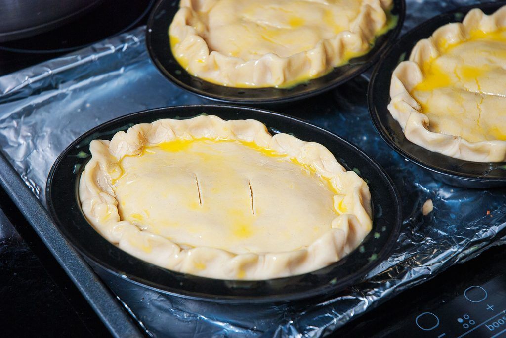 Mini Tourtiere meat pies brushed with beaten egg, with slits on top, arranged on a tray and ready to bake.
