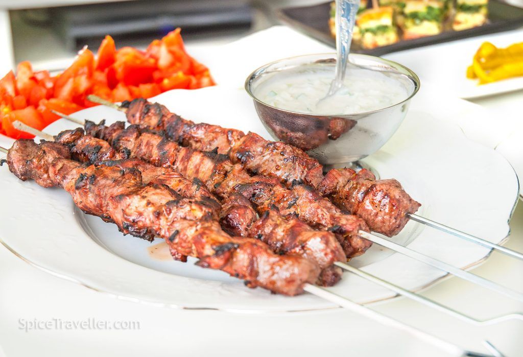 Grilled lamb skewers served on a plate, with a small silver bowl of yoghurt and cucumber sauce on the side.