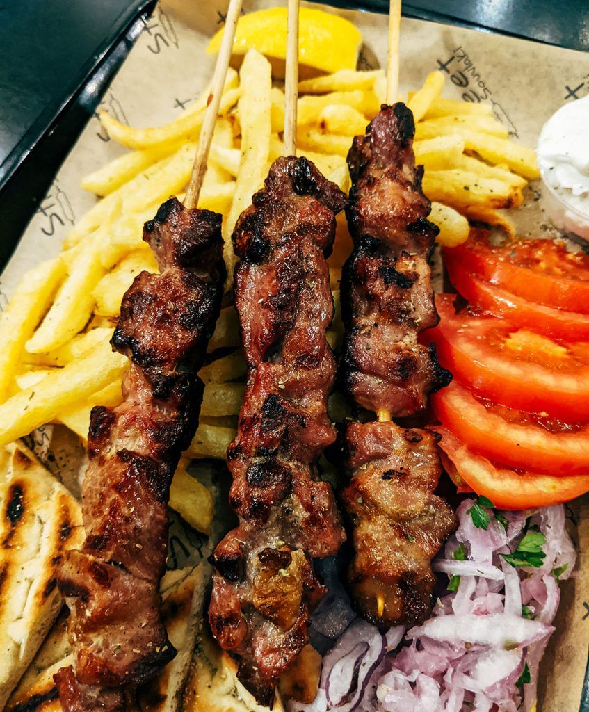 Grilled lamb skewers served with sliced tomatoes, warm pita bread, and a side of fries on a tray.