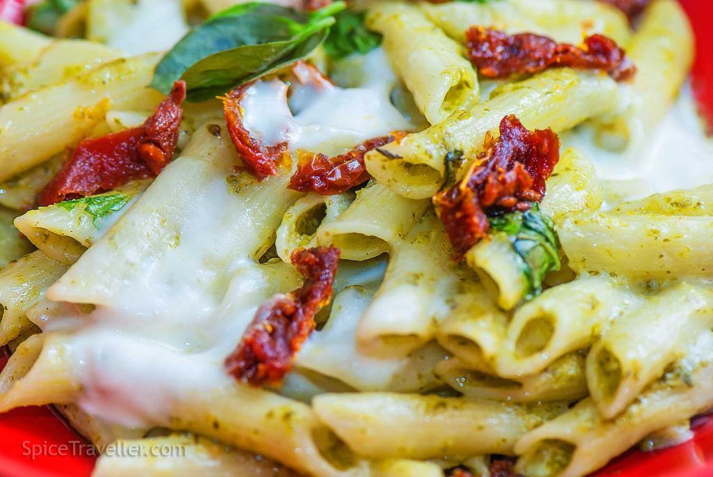 Close-up of green pesto penne pasta with melted buffalo mozzarella and pieces of sun-dried tomatoes.