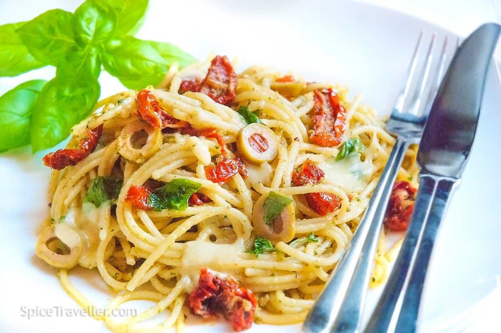 Serving suggestion: green pesto pasta with spaghetti, green olives stuffed with peppers, sun-dried tomatoes, breadcrumbs, and basil leaves on a white plate.