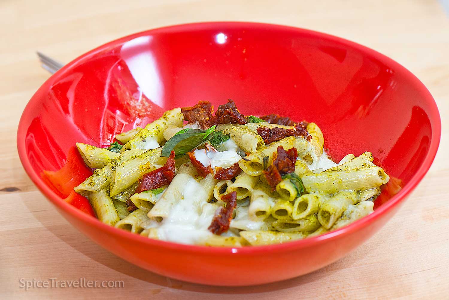 Pesto pasta with penne, black olives, sun-dried tomatoes, breadcrumbs, and fresh basil served in a red bowl.
